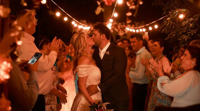 Couple shares a tender kiss surrounded by friends at a romantic outdoor wedding at night.